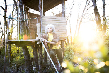 Ältere Frau lehnt sich bei Sonnenuntergang gegen die Leiter eines Baumhauses im Wald - CUF04583