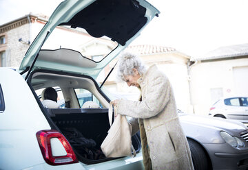 Ältere Frau mit Einkaufstasche schaut in den Kofferraum eines Autos - CUF04592