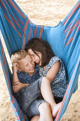 Girl and brother laughing huddled in hammock on Poetto beach, Cagliari, Italy - CUF04697