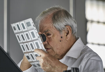 Senior man looking at sheet of film slides with magnifier - CUF04707