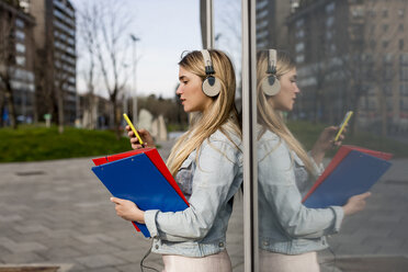 Junge Frau, im Freien, an ein Fenster gelehnt, mit Kopfhörern, Smartphone und Aktenordner in der Hand - CUF04713
