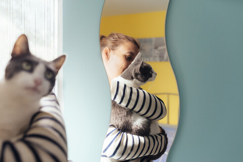 Frau hält eine graue Katze neben einem Spiegel zu Hause, lizenzfreies Stockfoto