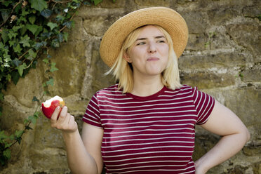 Porträt einer jungen blonden Frau mit Sonnenhut, die einen Apfel isst - FLLF00040