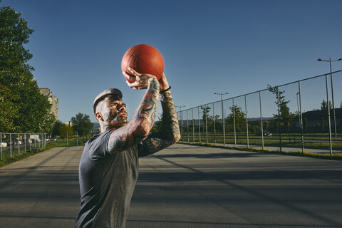 Tätowierter junger Mann wirft Basketball auf einem Platz im Freien, lizenzfreies Stockfoto
