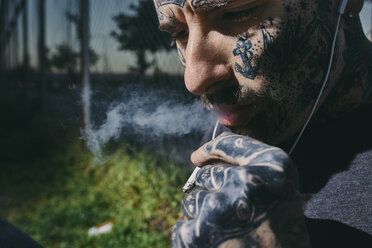 Nahaufnahme eines tätowierten jungen Mannes mit Ohrstöpseln, der eine Zigarette raucht - ZEDF01470