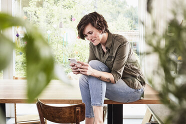 Mid adult woman at home, sitting on table, using smartphone - ISF01272