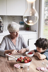 Grandmother sitting at kitchen table, preparing strawberries, grandson sitting beside her, watching - ISF01278