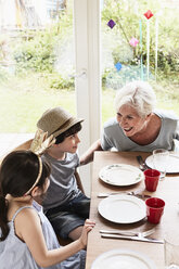 Grandmother sitting at kitchen table with grandson and granddaughter, smiling - ISF01286