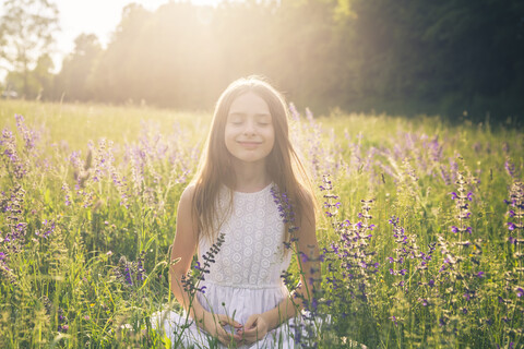 Portrait of smiling girl crouching on flower meadow at evening twilight stock photo