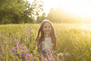 Porträt eines lächelnden Mädchens, das in der Abenddämmerung auf einer Blumenwiese hockt - SARF03747
