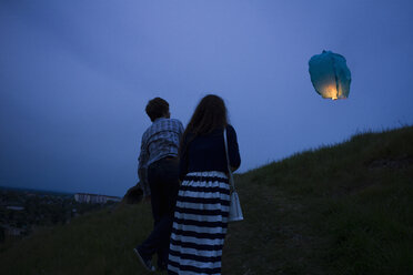 Couple walking up grassy hill at dusk, watching sky lantern in air, rear view - CUF04948