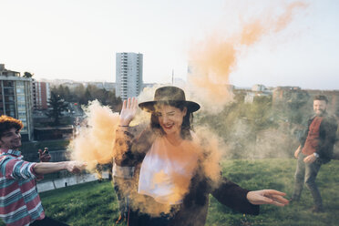 Group of friends on roof, holding colourful smoke flares, young woman walking through orange smoke - CUF05053