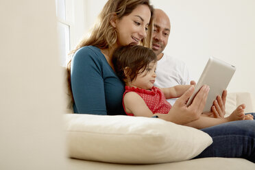 Ein kleines Mädchen sitzt mit seinen Eltern auf dem Sofa und schaut auf ein digitales Tablet - CUF05093