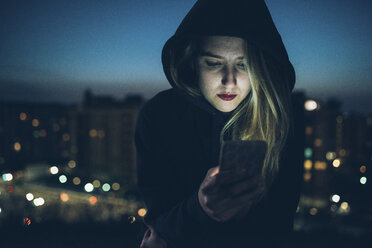 Junge Frau sitzt nachts auf einem Dach und benutzt ein Smartphone, das das Gesicht beleuchtet - CUF05121
