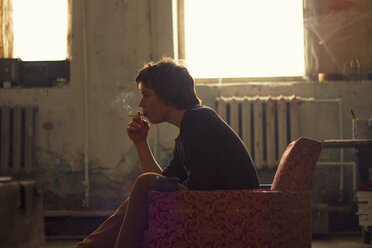 Junger Mann sitzt auf einem alten Sessel im Künstleratelier und raucht eine Zigarette - CUF05160