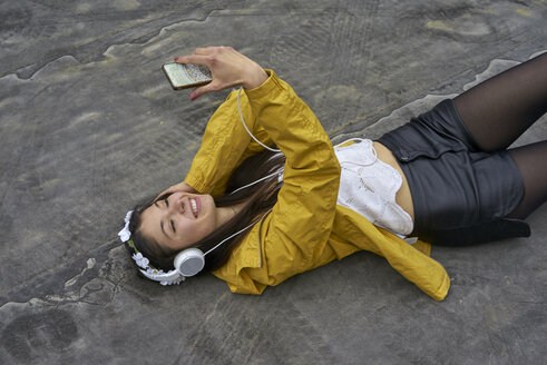 Lächelnde Frau mit Kopfhörern, die auf dem Boden liegt und ein Selfie mit ihrem Smartphone macht - BEF00049