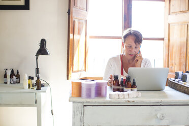 Frau, die mit ihrem Smartphone spricht, während sie einen Laptop im Büro einer Werkstatt für handgemachte Seife benutzt - CUF05441