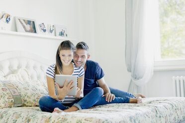 Pregnant couple sitting on bed using digital tablet smiling - CUF05456