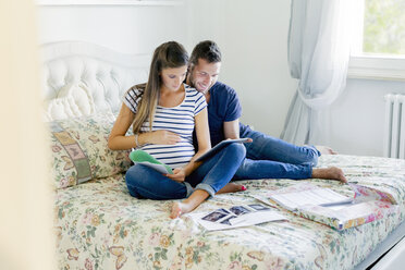 Pregnant couple sitting on bed using digital tablet smiling - CUF05457