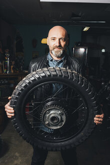 Porträt eines reifen Mannes in einer Garage, der einen Motorradreifen hält - CUF05693