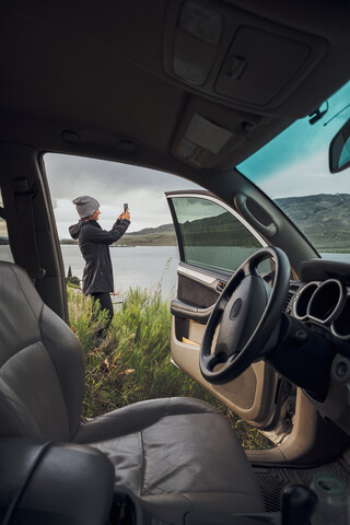 Junge Frau steht am Dillon Reservoir und hält ihr Smartphone, Blick durch ein geparktes Auto, Silverthorne, Colorado, USA, lizenzfreies Stockfoto