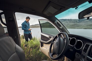 Mittlerer erwachsener Mann steht am Dillon Reservoir und hält ein Smartphone, Blick durch ein geparktes Auto, Silverthorne, Colorado, USA - ISF01361