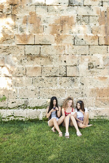 Drei junge Frauen sitzen an einer Steinmauer und hören mit ihren Handys Musik - IGGF00479