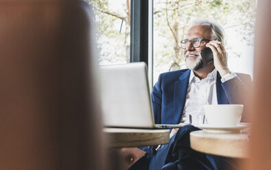 Mature businessman sitting at table in a cafe using cell phone and laptop - UUF13677