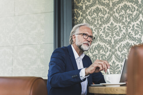 Reifer Geschäftsmann sitzt mit Laptop und Tasse Kaffee am Tisch in einem Café - UUF13679