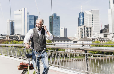 Smiling mature man with earbuds and bicycle crossing bridge in the city - UUF13702