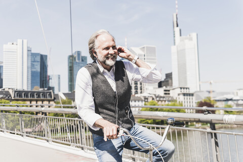 Smiling mature man with earbuds on bicycle crossing bridge in the city stock photo