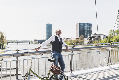 Mature man with bicycle listening to music on bridge in the city - UUF13708