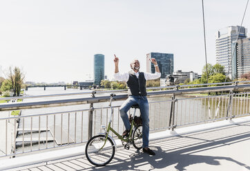 Glücklicher reifer Mann mit Fahrrad hört Musik auf einer Brücke in der Stadt - UUF13709