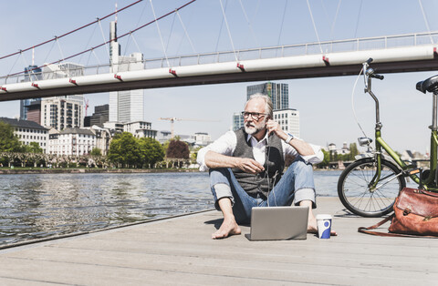 Mature man with laptop, earbuds and bicycle sitting at the riverside in the city stock photo