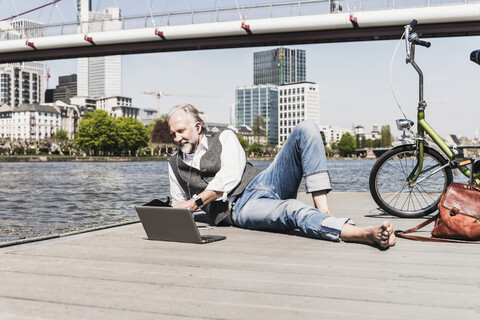 Älterer Mann mit Laptop, Kopfhörern und Fahrrad am Flussufer in der Stadt liegend, lizenzfreies Stockfoto