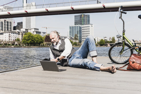 Mature man with laptop, earbuds and bicycle lying at the riverside in the city - UUF13711