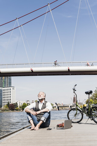 Mature man with laptop and bicycle sitting at the riverside in the city stock photo