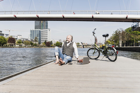 Lächelnder reifer Mann mit Laptop und Fahrrad am Flussufer in der Stadt sitzend, lizenzfreies Stockfoto