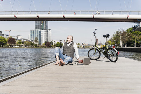 Smiling mature man with laptop and bicycle sitting at the riverside in the city - UUF13715