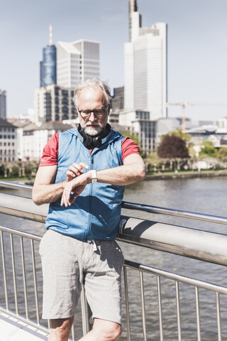 Sportlicher reifer Mann mit Kopfhörern, der sich auf einer Brücke in der Stadt die Zeit nimmt, lizenzfreies Stockfoto