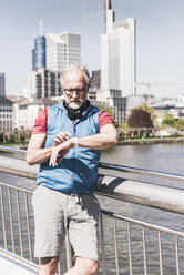 Sportlicher reifer Mann mit Kopfhörern, der sich auf einer Brücke in der Stadt die Zeit nimmt - UUF13721