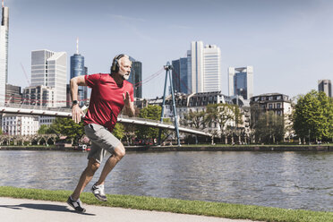 Mature man with headphones running at the riverside in the city - UUF13723