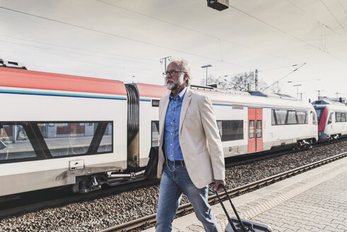 Älterer Geschäftsmann mit Ohrstöpseln und Koffer auf dem Bahnsteig - UUF13732