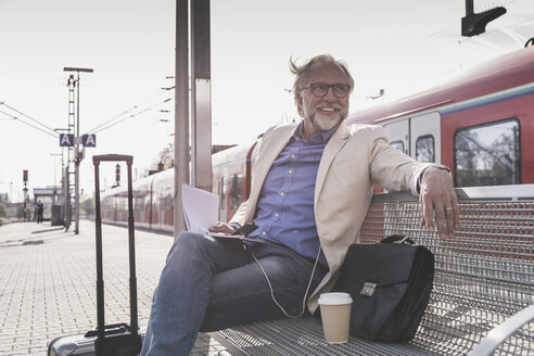 Lächelnder reifer Geschäftsmann sitzt am Bahnhof mit Handy, Kopfhörern und Notebook - UUF13734