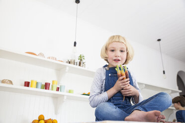 Porträt eines süßen Mädchens, das auf der Küchentheke sitzt und ein Bündel bunter Karotten hält - CUF05724