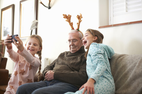 Sisters taking smartphone selfie of sleeping grandfather in reindeer antlers - CUF05745