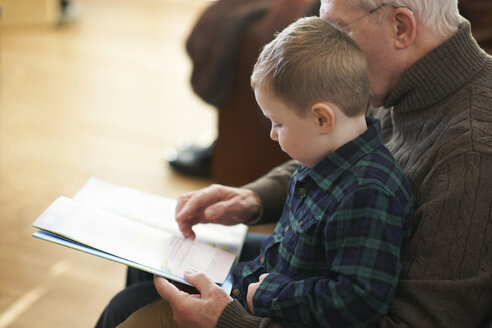 Junge sitzt auf Großvaters Schoß und liest ein Buch - CUF05750