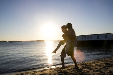 Beleuchteter junger Mann, der seine Freundin am Strand bei Sonnenuntergang Huckepack nimmt - CUF05886