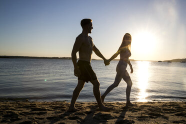 Beleuchteter junger Mann und seine Freundin spazieren Hand in Hand am Strand bei Sonnenuntergang - CUF05887