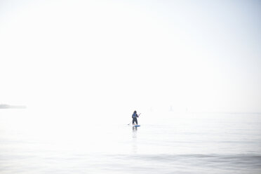 Distant view of female paddleboarder paddling out on misty sea - CUF05953